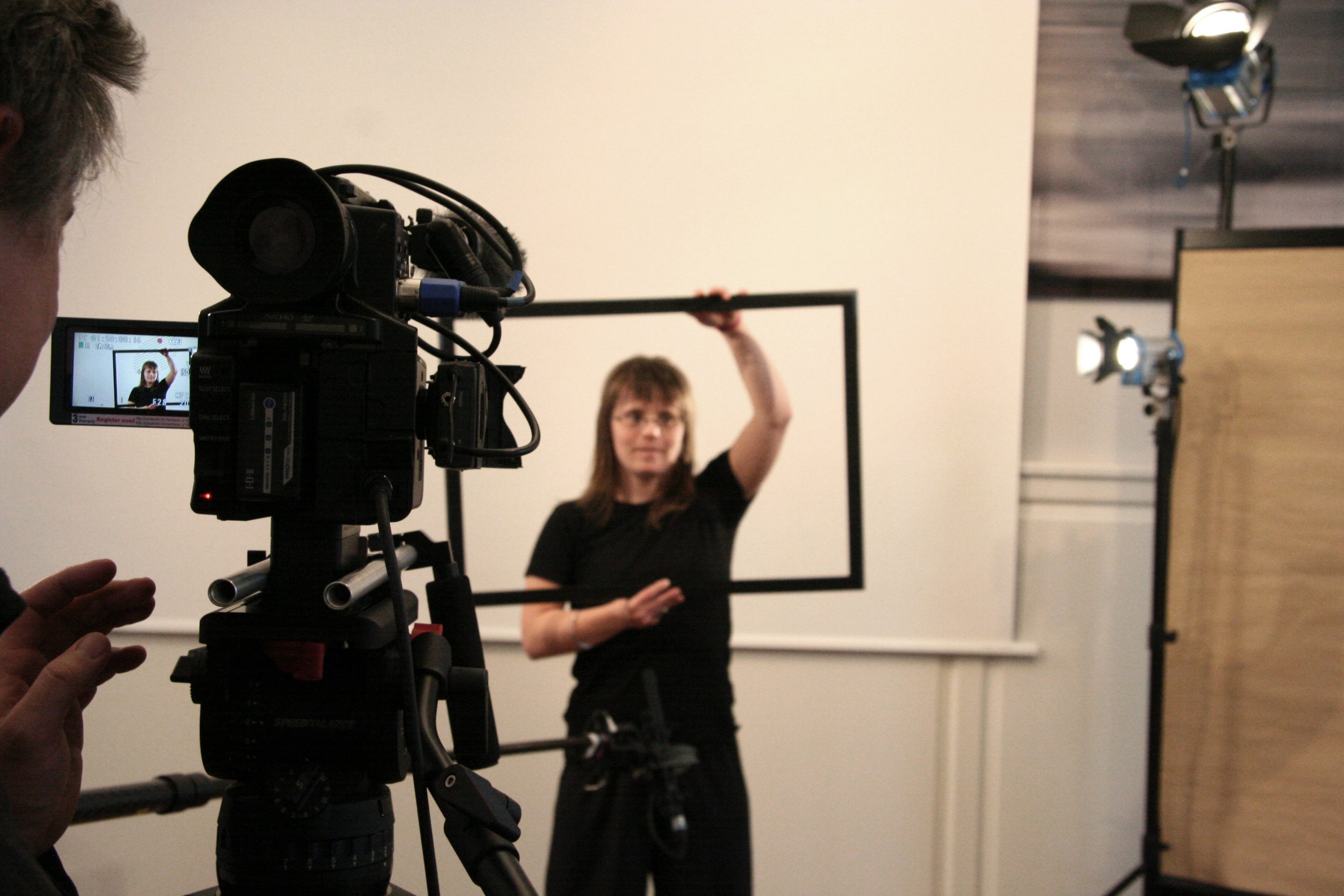 Eine Frau hält einen Bilderrahmen vor ihrem Oberkörper und steht vor einer Videokamera.