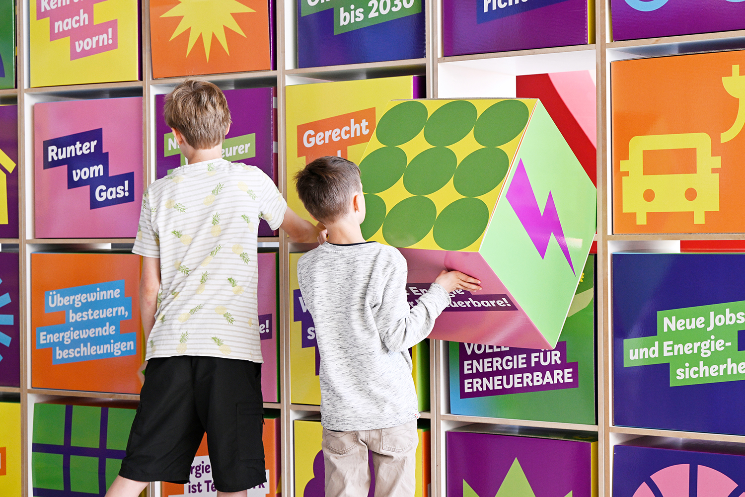 Zwei Jungen stehen vor einer bunten Wand bestehend aus Würfeln mit verschiedenen Symbolen und Texten.