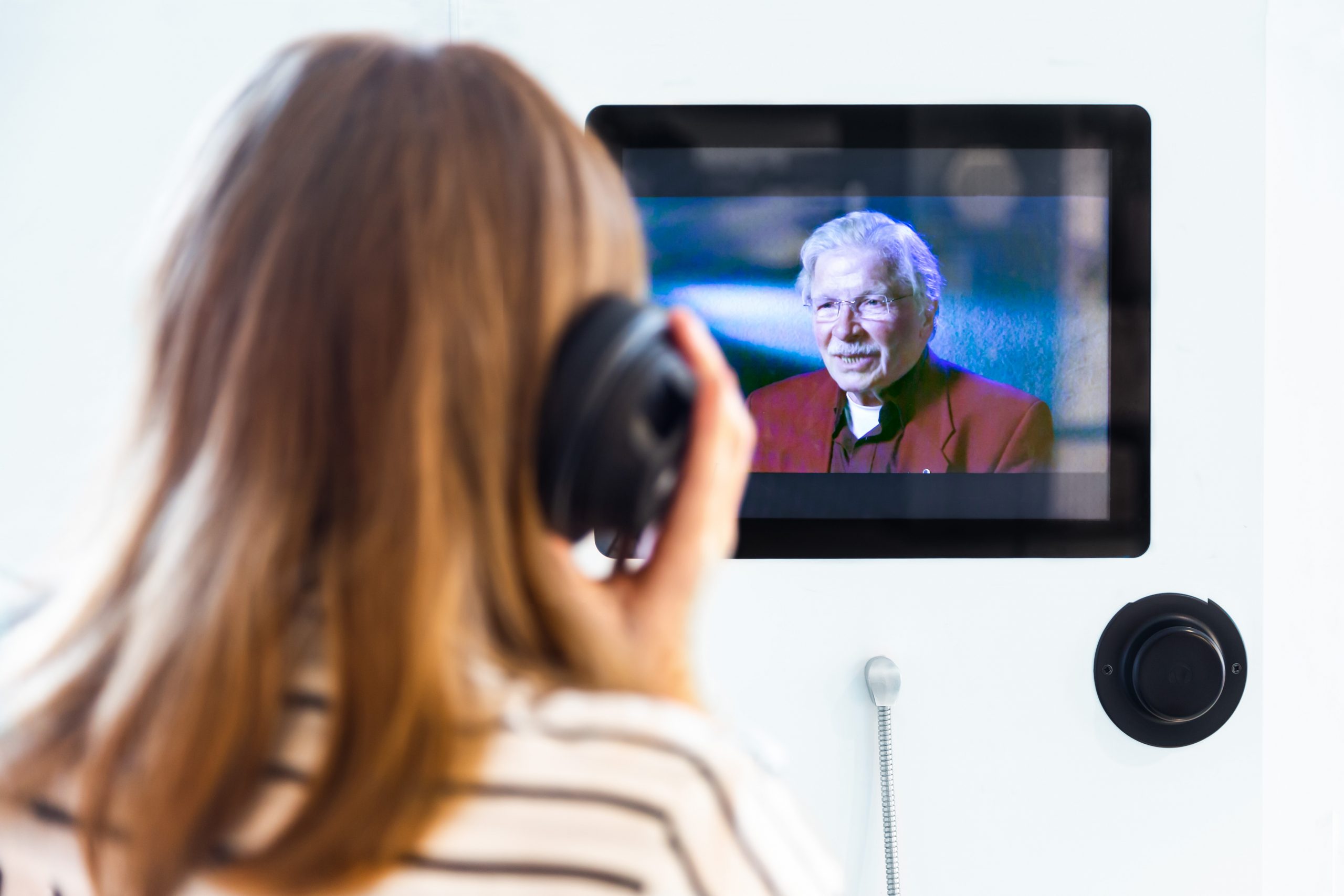 Eine Frau, die man von hinten sieht, sitzt mit einem Kopfhörer vor einem Bildschirm.