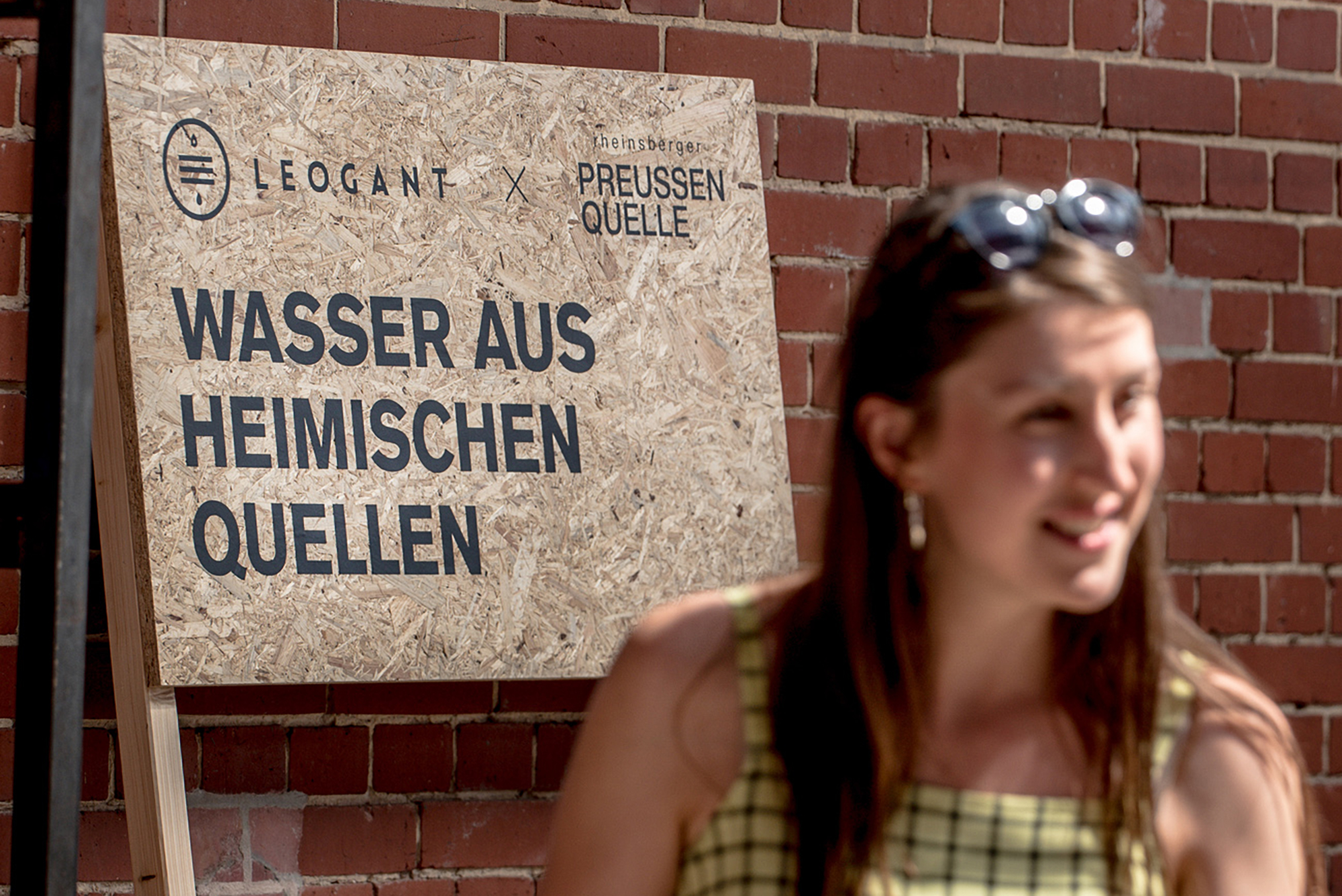 Eine Frau an der Wasserbar steht vor einem Schild mit den Logos von Leogant und Rheinsberger Preussenquelle und dem Schriftzug: Wasser aus heimischen Quellen.