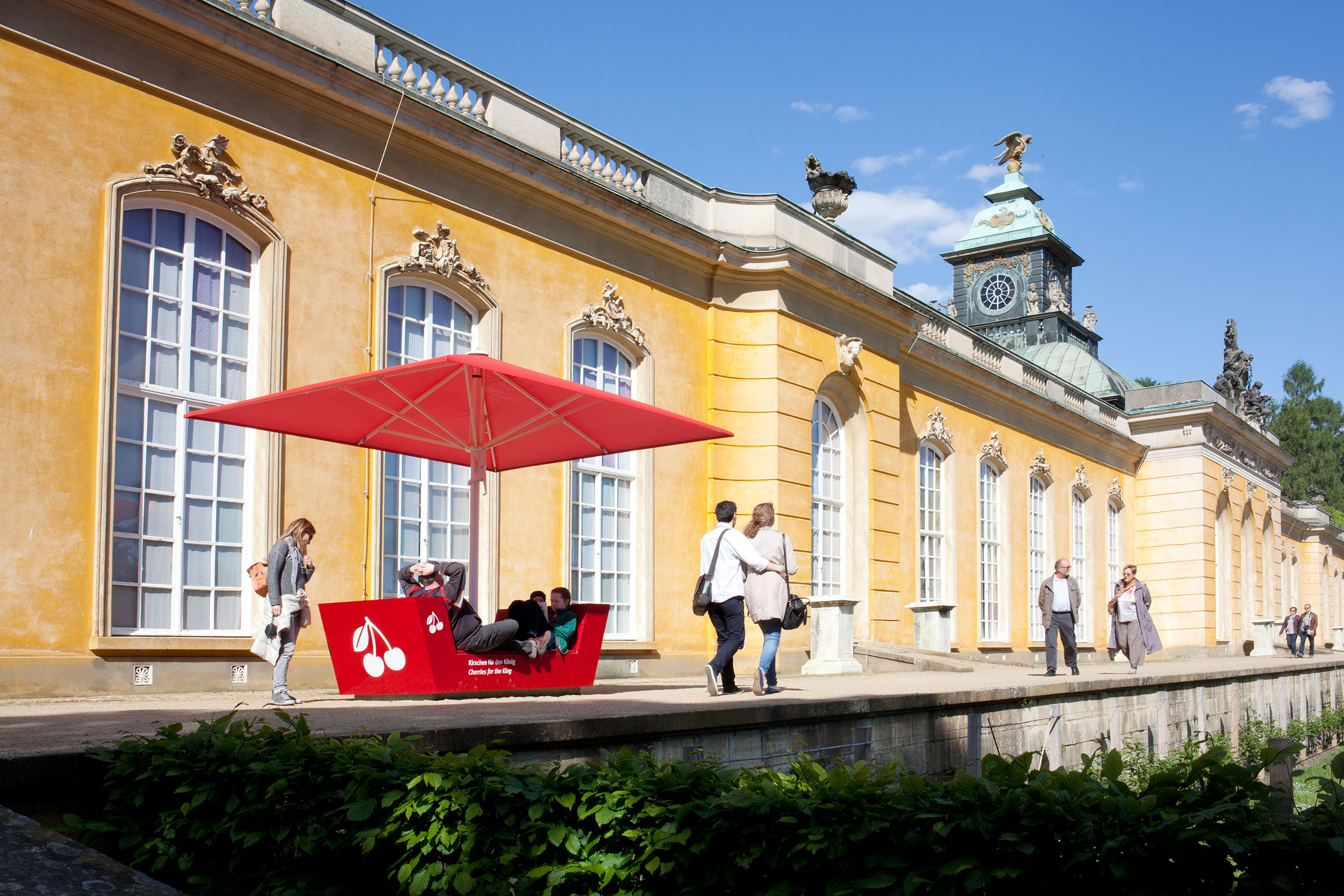 Vor dem Schloss Sanssouci: Eine rote Informationsstation der Outdoorausstellung Paradiesapfel, die gleichzeitig Sitzgelegenheit inklusive Sonnenschirm ist.