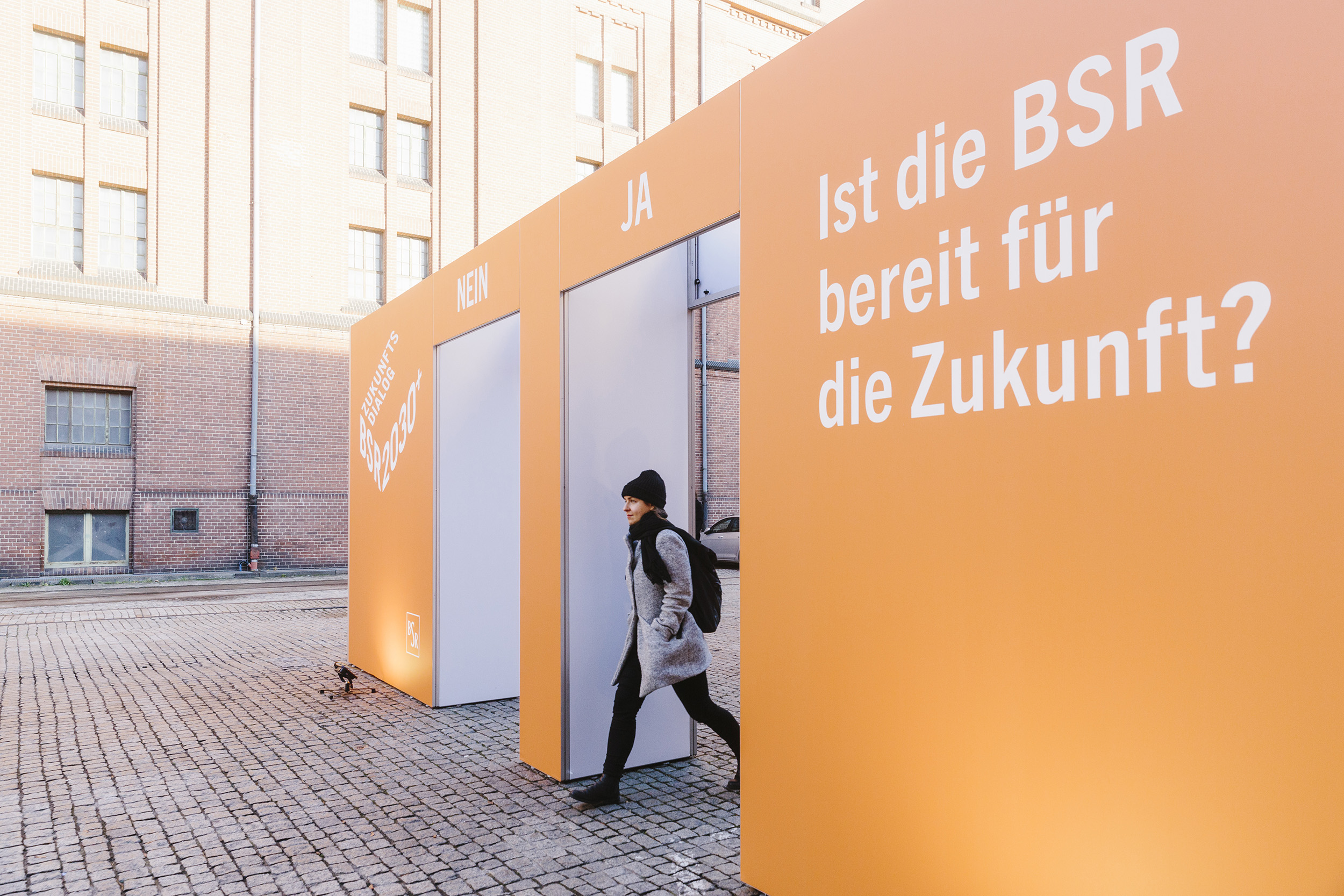 Eine Frau, die durch ein großes orangefarbenen Tor durch geht. Auf dem Tor steht die Frage: "Ist die BSR bereit für die Zukunft?".