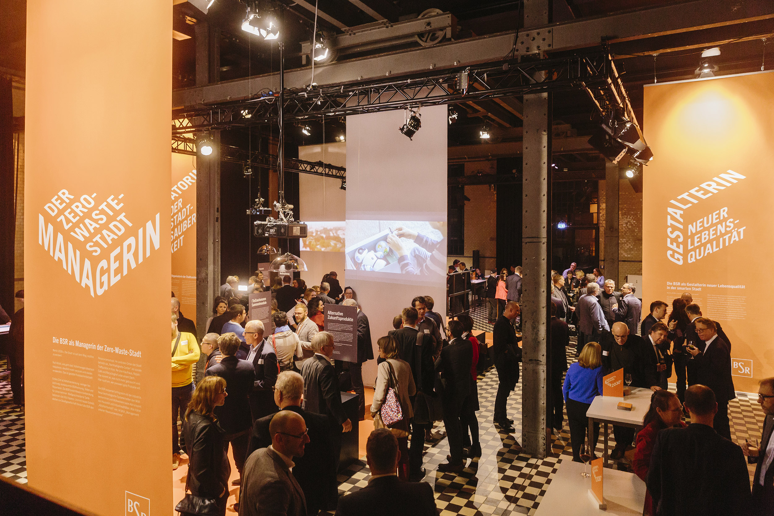 Eine Gruppe an Menschen in einer Halle, umgeben von orange-farbigen Ausstellungsinseln.