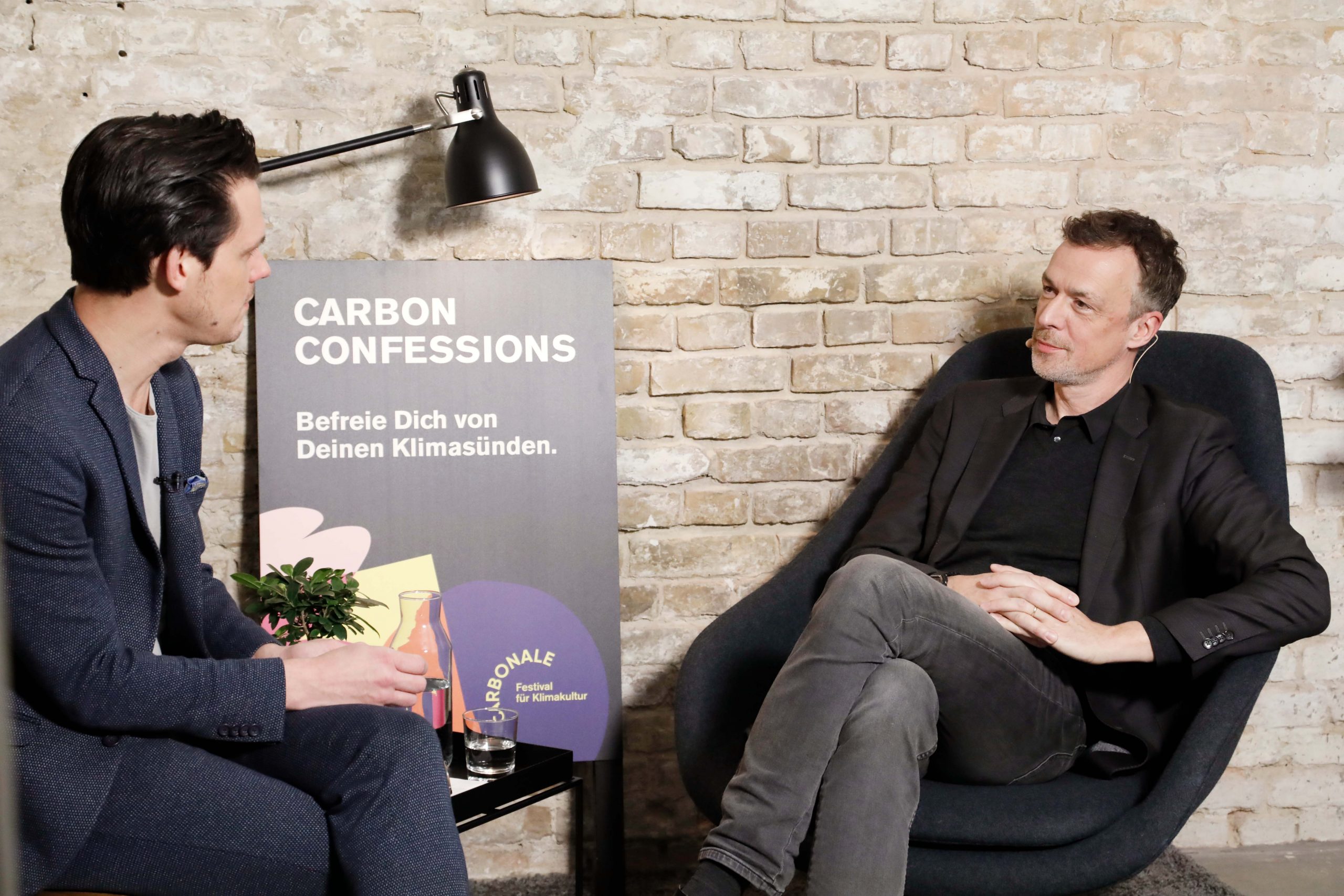 Carbon Confessions – Ein Mann, der auf einem Stuhl sitzt und mit einem anderen Mann spricht.