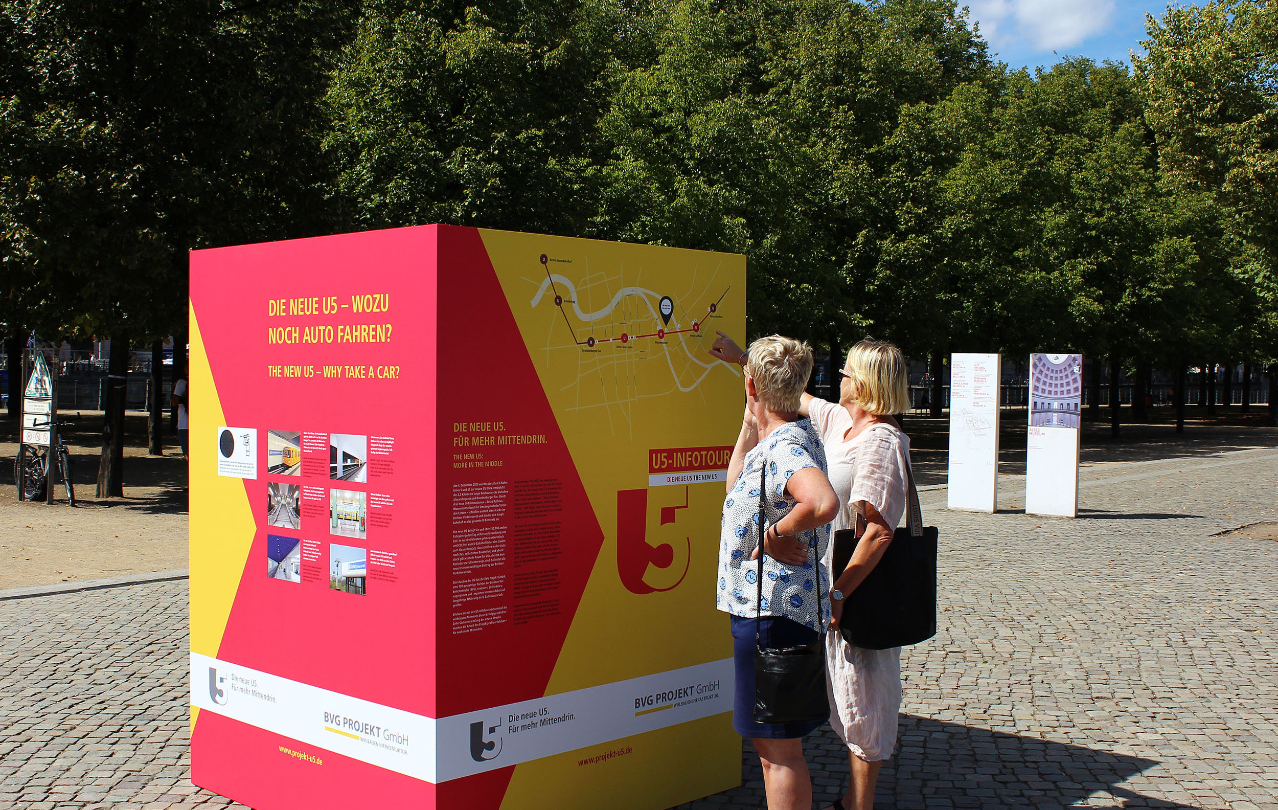 Zwei Frauen stehen vor einer rot-gelben Information der U5 Infotour und schauen sich eine Karte an.