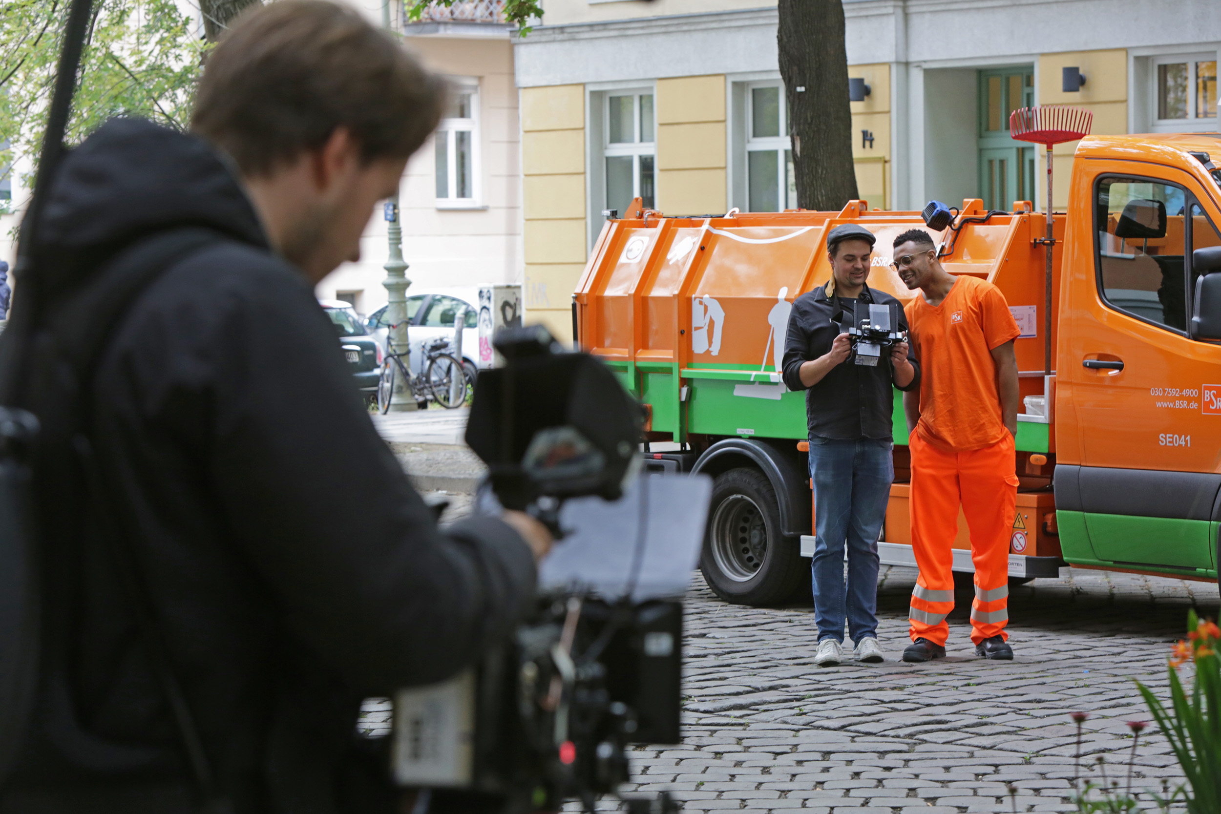 Ein Kameramann filmt zwei Männer vor einem BSR Auto. Einer davon trägt orangene Arbeitskleidung.